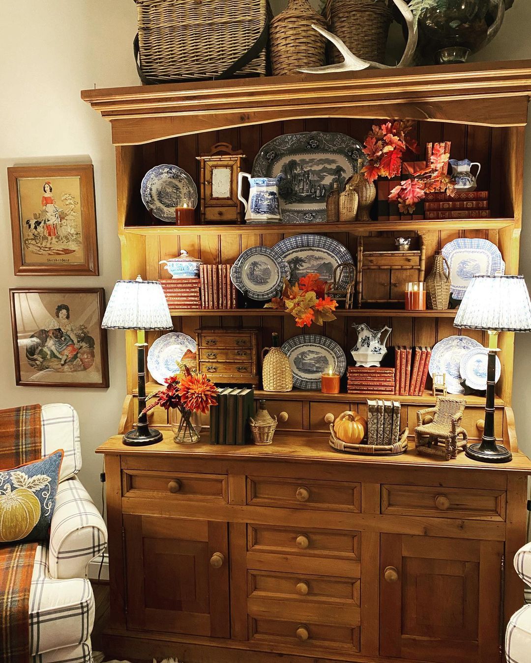 Pine hutch styled for autumn with blue and white transferware plates red cloth books autumn foliage amber candles and two pleated lampshades on the surface
