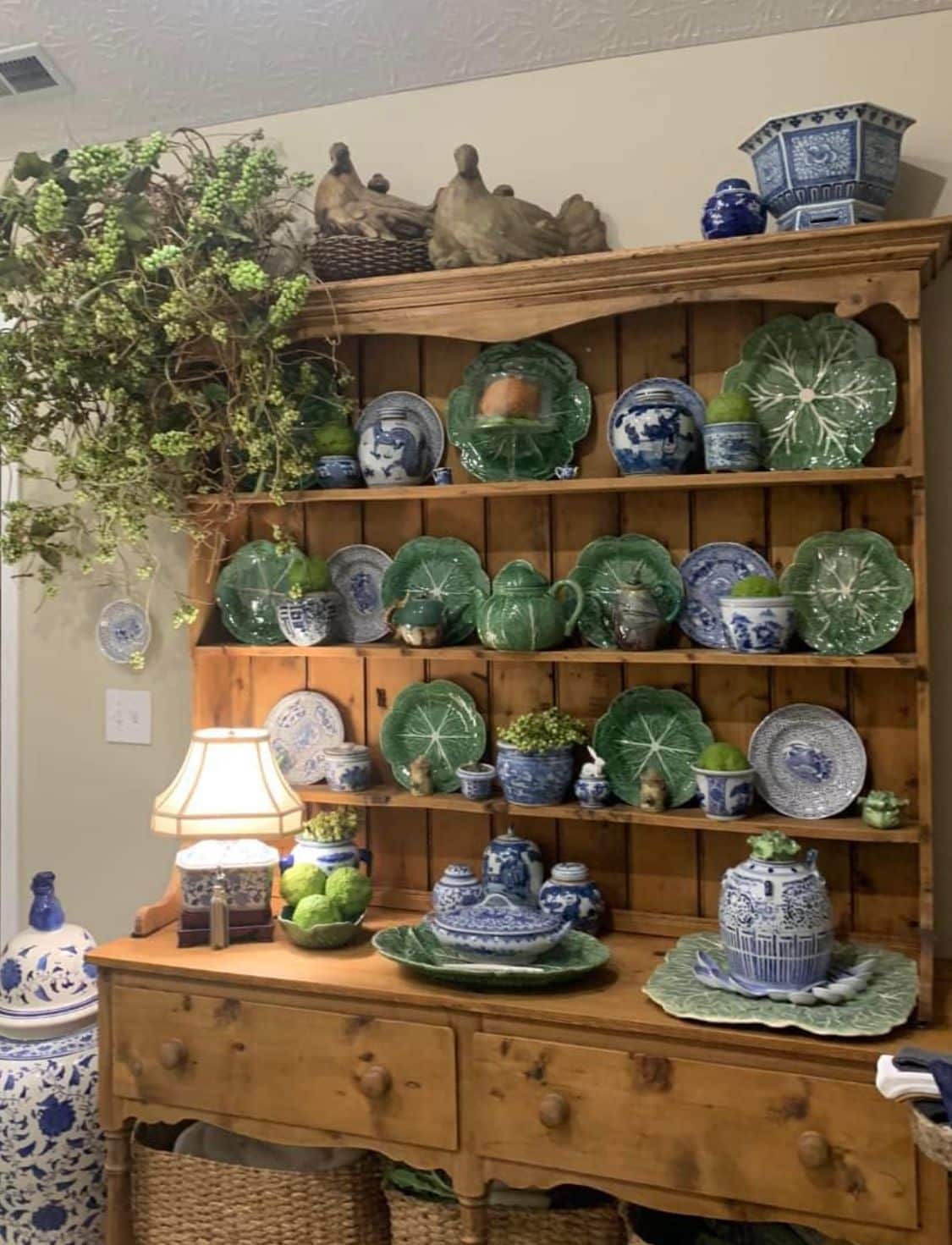 Pine hutch with green majolica leaf plates blue and white ginger jars and transferware plates on every shelf with trailing greenery and a lamp on the surface