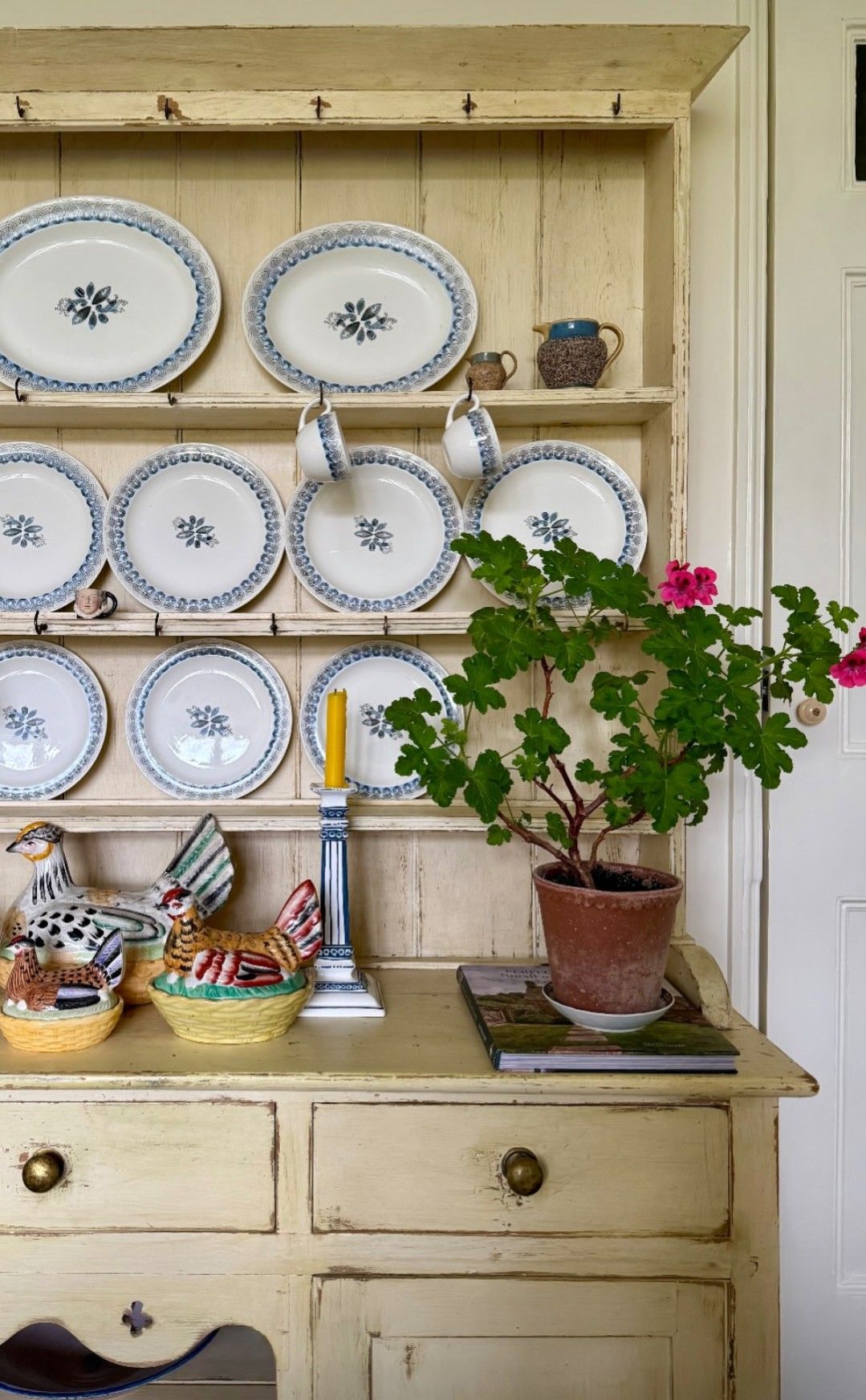 Cream painted pine hutch with matching blue and white oval plates displayed on every shelf a yellow candle blue candlestick ceramic hen figures and a potted geranium