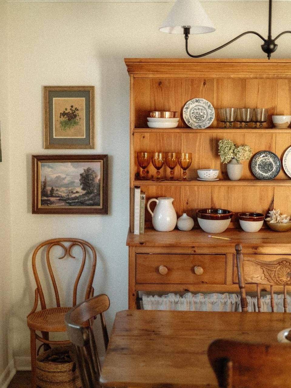 Pine hutch in a dining room with amber glass goblets white pitcher stoneware mixing bowls and vintage plates on shelves beside framed landscape paintings