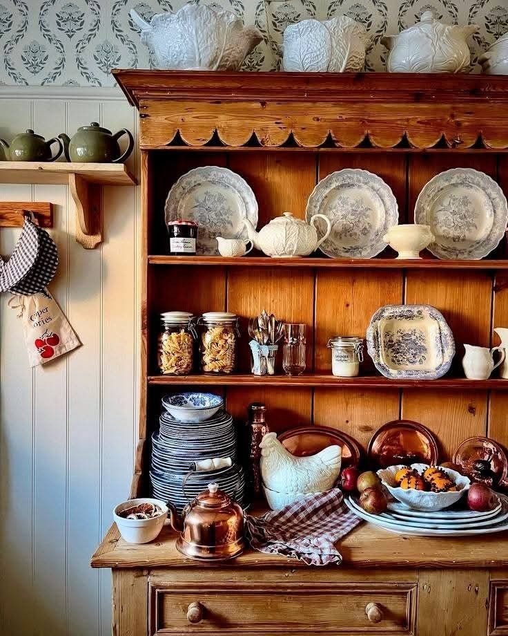 Pine hutch with all-white ironstone plates teapot and serving pieces with copper kettle and gingham linen on the surface against patterned wallpaper