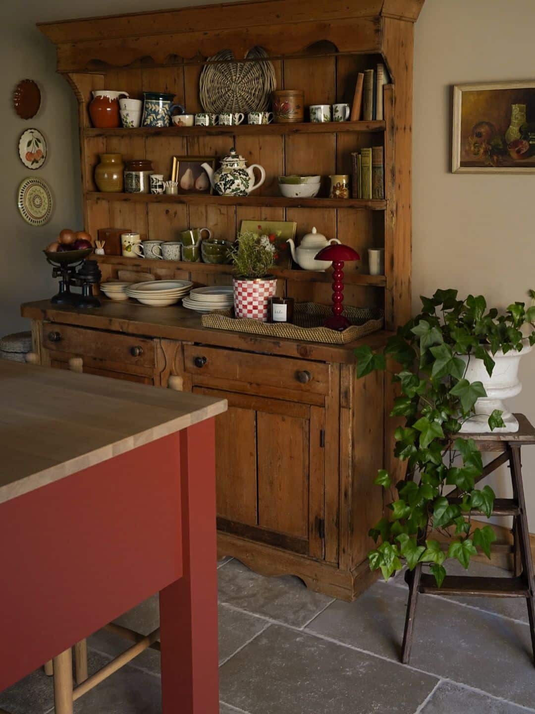 Dark pine hutch in a farmhouse kitchen with pottery mugs teapots and jugs on shelves a rattan tray vignette with a red lamp and trailing ivy nearby