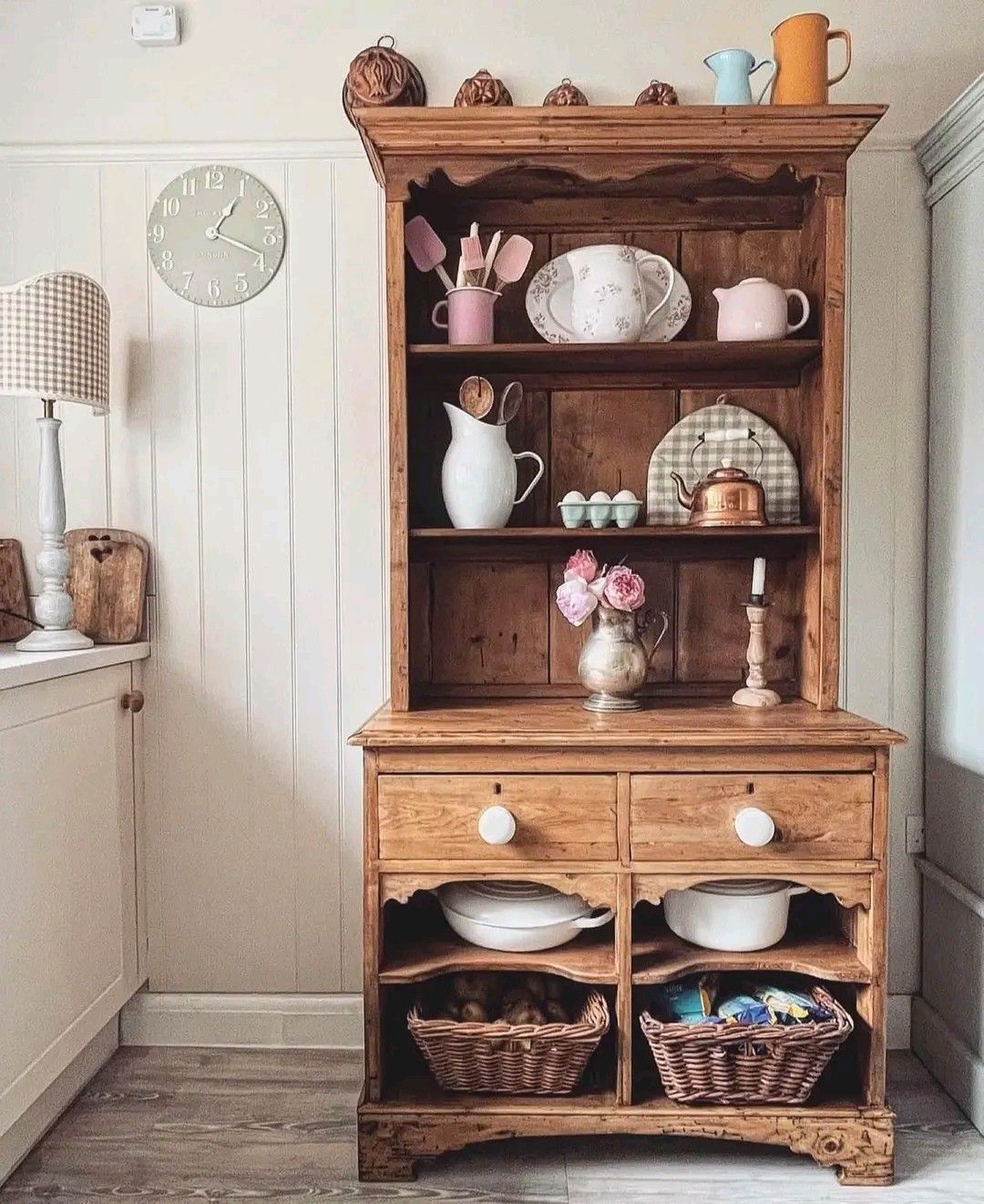 Pine hutch in a cottage kitchen with pink kitchen utensils white jug fresh pink roses copper kettle and wicker baskets storing cast iron pots below
