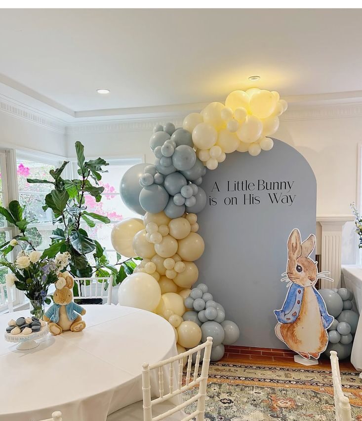 Peter Rabbit baby shower arch panel backdrop in dusty blue with a cream and blue balloon garland a Little Bunny is on His Way text and a Peter Rabbit character cutout