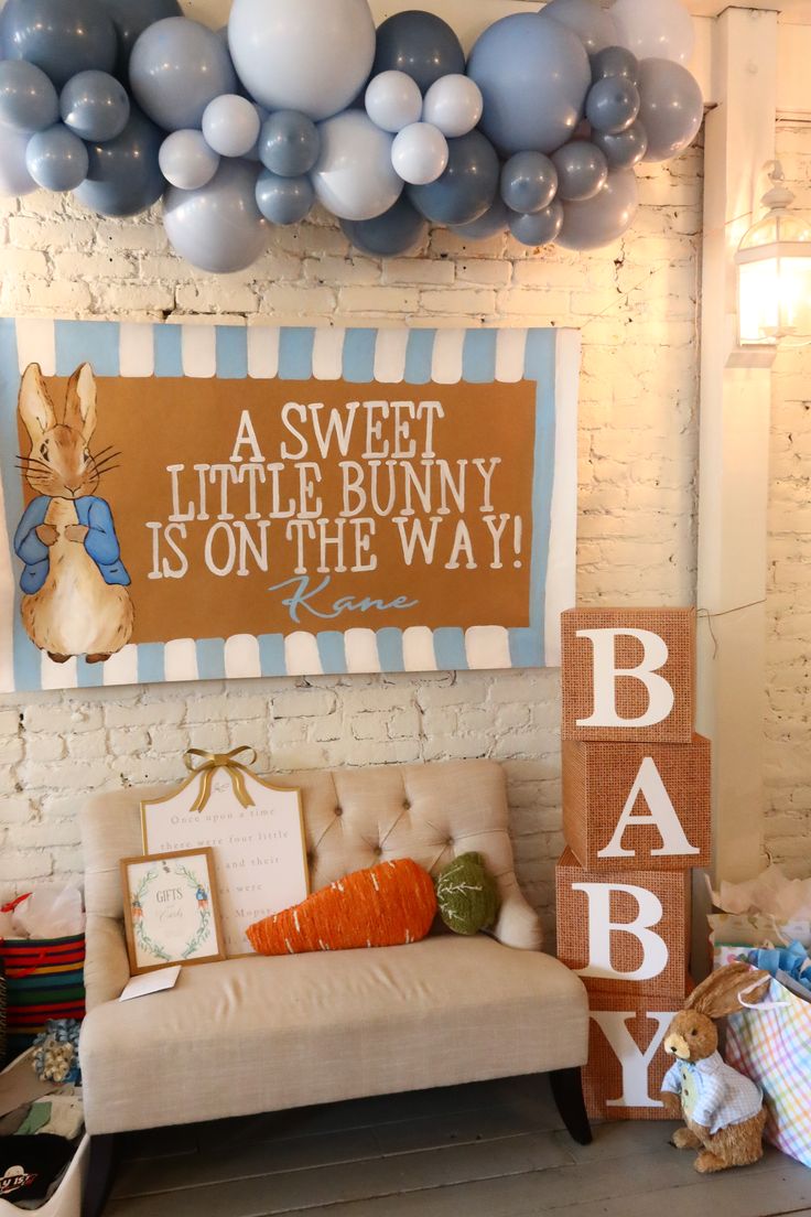 Peter Rabbit baby shower personalised banner reading A Sweet Little Bunny is on the Way with a Peter Rabbit illustration on a white brick wall with burlap BABY blocks and blue balloon garland
