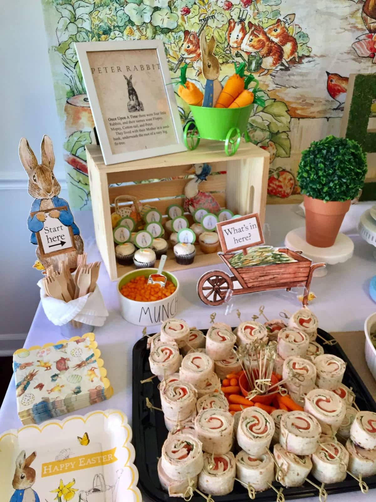 Peter Rabbit baby shower food table with sandwich pinwheels cupcakes in a wooden crate a mini wheelbarrow with fabric carrots character cutouts and a framed storybook excerpt