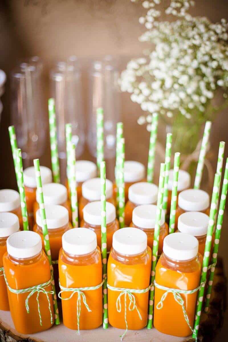Peter Rabbit baby shower individual carrot juice bottles tied with green twine and topped with green polka dot paper straws displayed on a wooden slice