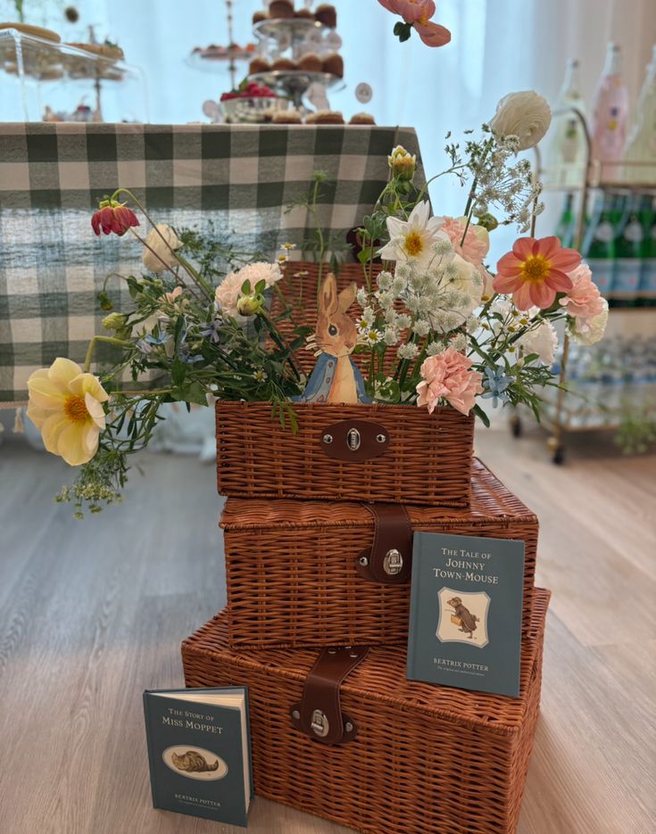 Stacked wicker hamper baskets with wildflowers dahlias carnations and daisies spilling from the top and Beatrix Potter books displayed at the base
