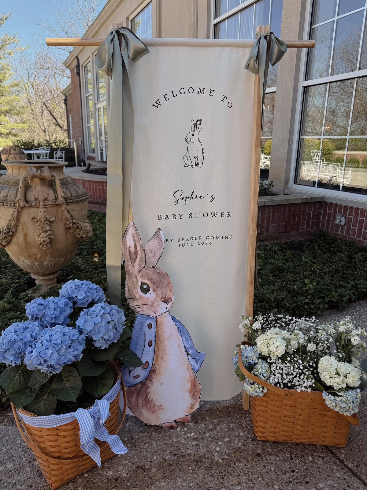 Personalised linen Peter Rabbit baby shower welcome banner on a timber frame with sage ribbon bows flanked by wicker baskets of blue hydrangeas and a Peter Rabbit cutout