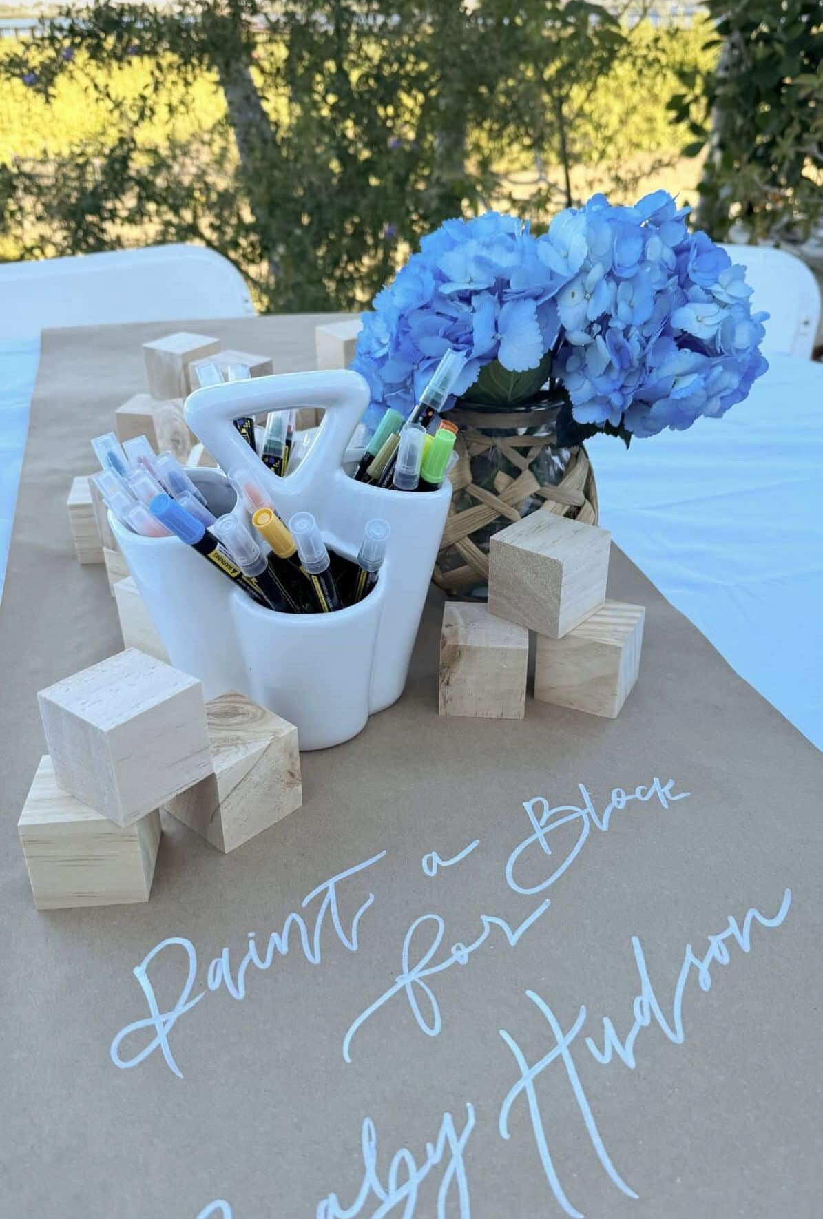 Peter Rabbit baby shower paint a block activity station with plain wooden blocks and markers on a kraft paper covered table with blue hydrangeas in a wicker vase