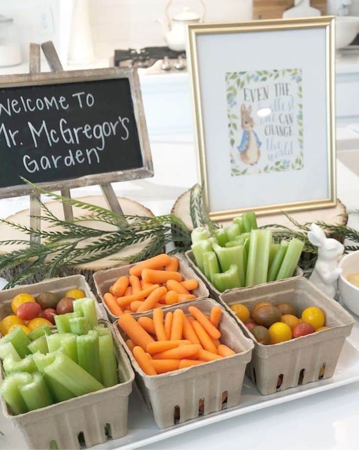 Peter Rabbit baby shower snack station with cardboard produce boxes filled with carrots celery and cherry tomatoes labelled Welcome to Mr McGregor's Garden with a framed print
