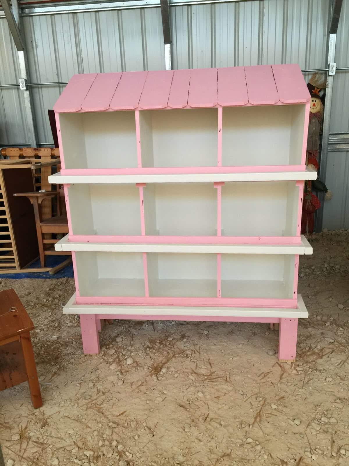 A freestanding nine-compartment wooden nesting box unit painted pink and cream with a decorative scalloped roof, three rows of three open compartments, and raised legs, photographed in a barn