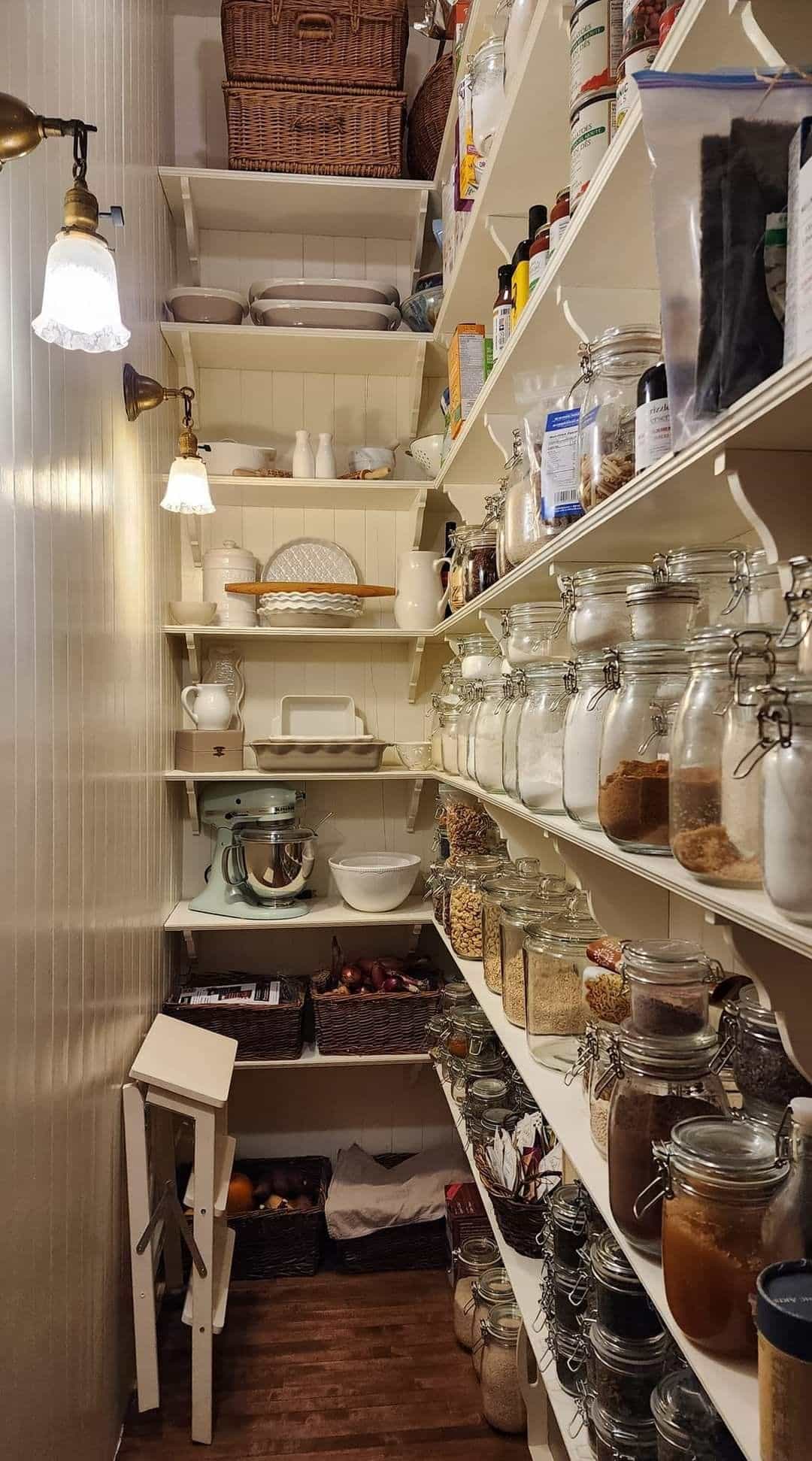 Walk-in farmhouse pantry with floor-to-ceiling white open shelves filled with glass clip-top jars containing dry goods, wicker baskets on upper shelves, brass vintage wall sconces, and a mint green KitchenAid stand mixer on a lower shelf