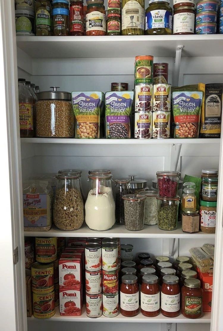 Simple white pantry shelves stocked with neatly organised canned tomatoes, pouched legumes, glass jars of oats and grains, pasta sauces, and other dry staples grouped together by type on each shelf