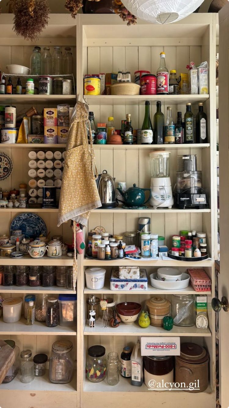 Tall cream freestanding farmhouse pantry cabinet with beadboard back panel and open shelves holding teas, oil bottles, kitchen appliances including a blender and food processor, spice jars, glass storage jars, and a yellow floral apron on a hook