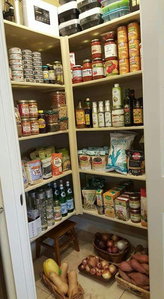 Corner walk-in pantry with cream L-shaped shelves stocked with oils, sauces, snacks, canned goods, and packaged foods, a small wooden step stool, and wicker floor baskets holding onions, sweet potatoes, and butternut squash