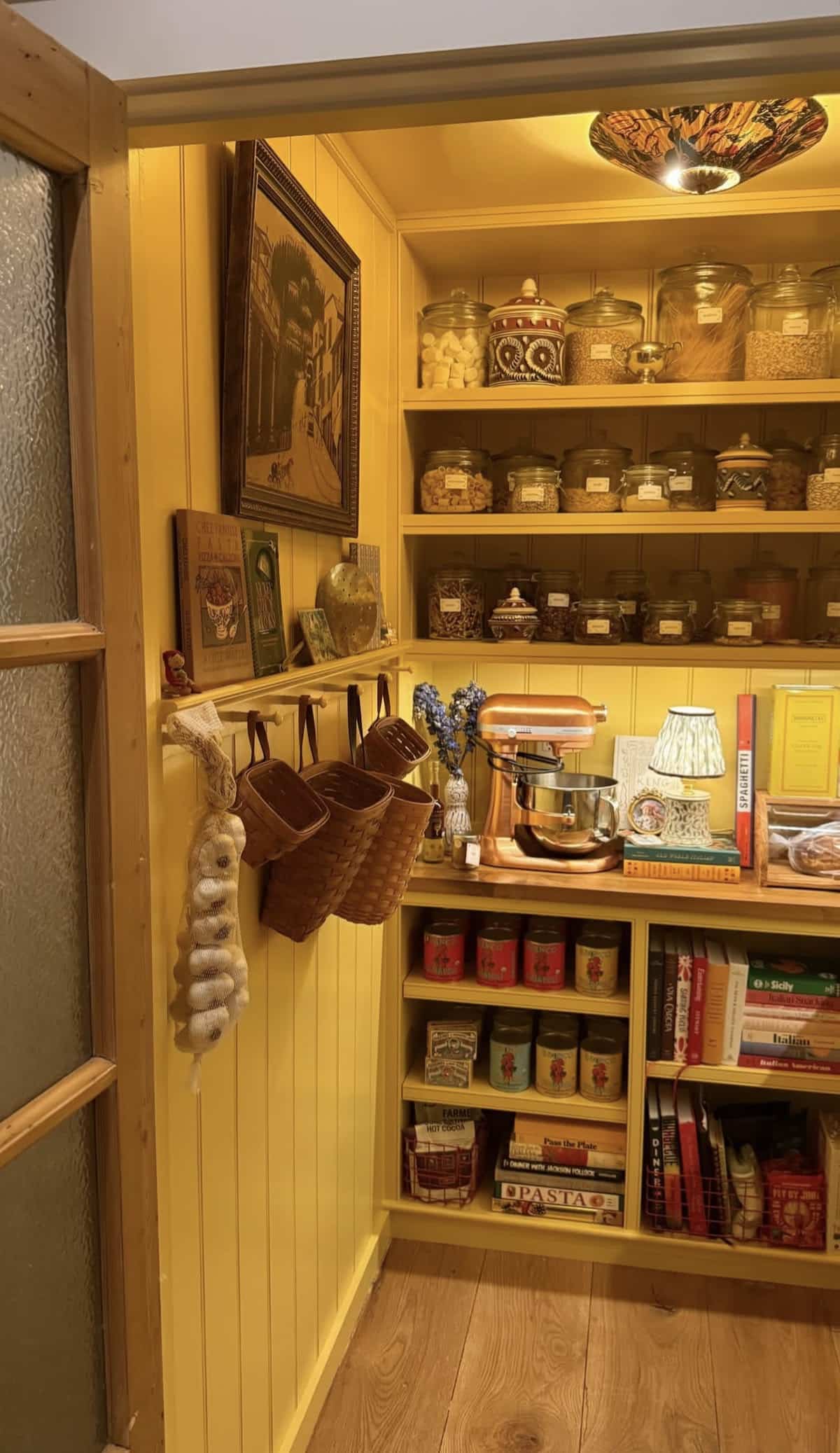 Warm golden yellow farmhouse pantry room with beadboard walls, tall open shelves of glass canisters, a butcher block work surface with a copper KitchenAid stand mixer, hanging market baskets on a wall rail, a Tiffany ceiling light, and a framed oil painting