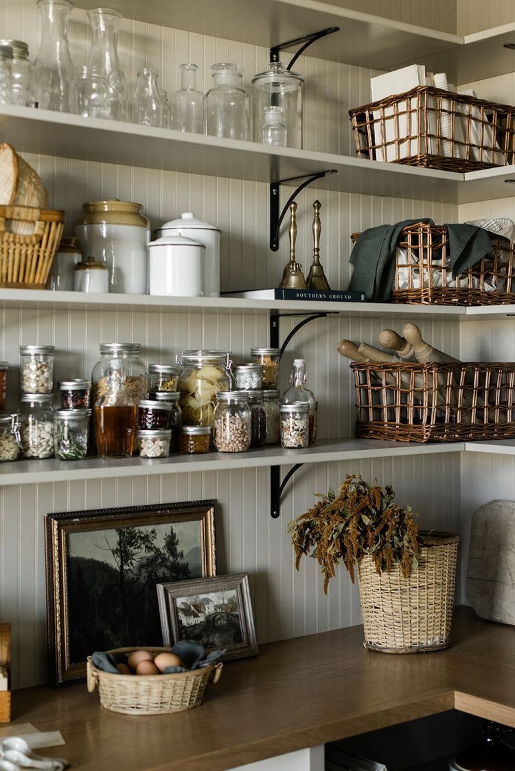 Farmhouse pantry shelves with greige beadboard backing and black iron brackets, holding glass jars, white enamel canisters, wicker baskets in two styles, a wooden bell, folded linen cloths, a basket of eggs, a framed vintage landscape, and dried botanicals
