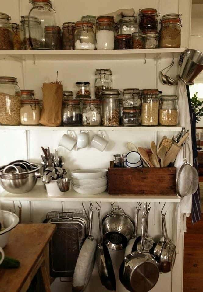 Open white farmhouse pantry shelves with clip-top glass jars for dry goods and spices, white enamel mugs on hooks, stacked mixing bowls, a wooden crate of kitchen utensils, and a lower hanging rail with stainless steel pots, pans, and a colander
