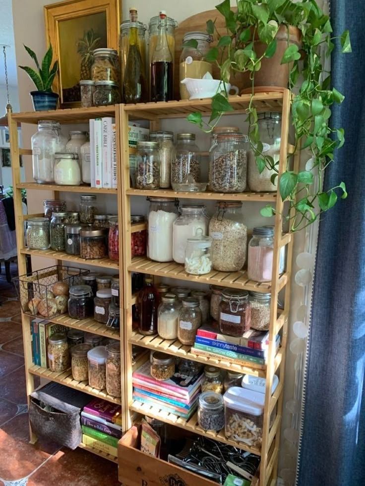 Two pine freestanding shelving units pushed together to form a pantry wall, filled with mason jars, swing-top glass jars, cookbooks, a wire basket of onions, ferment jars, and green trailing and potted houseplants growing across the top and side