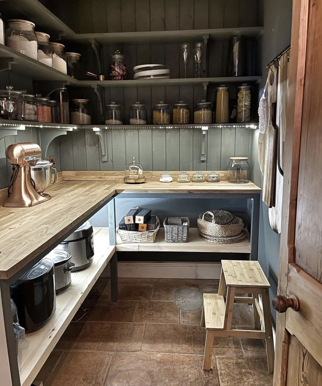 Moody dark slate blue tongue-and-groove farmhouse pantry room with wrap-around butcher block counters, LED strip shelf lighting, large glass storage jars on upper open shelves, a copper KitchenAid mixer, wicker baskets, and an original wooden door with a linen curtain