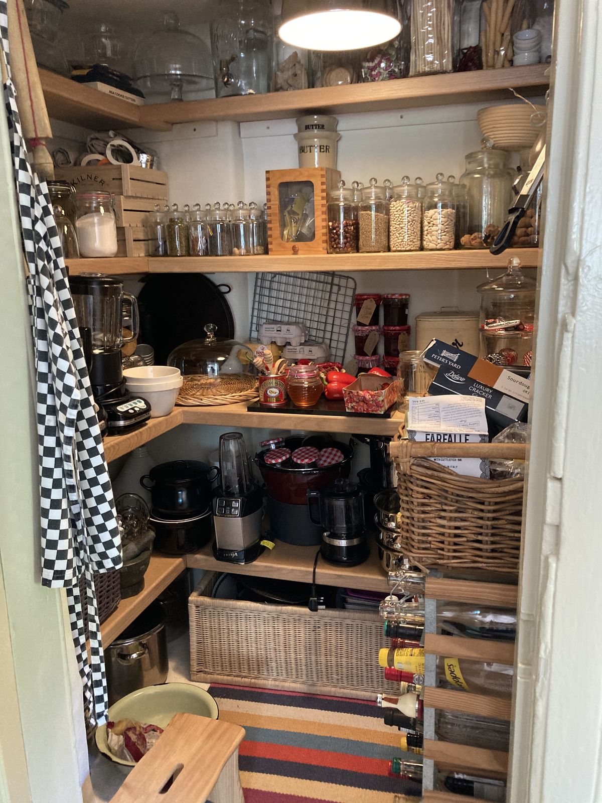 Compact corner farmhouse pantry with light oak shelves holding glass storage jars on upper levels, a blender, slow cooker, coffee maker, ceramic butter crock, wicker basket of pantry goods, a wall spice rack, a hanging checkered apron, and a striped floor rug