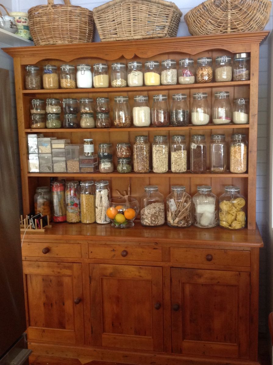 Antique pine farmhouse dresser with open hutch shelves filled front to back with Kilner swing-top glass jars of grains, seeds, and dried goods up to the decorative carved crown, large glass canisters on the counter, and wicker baskets on top