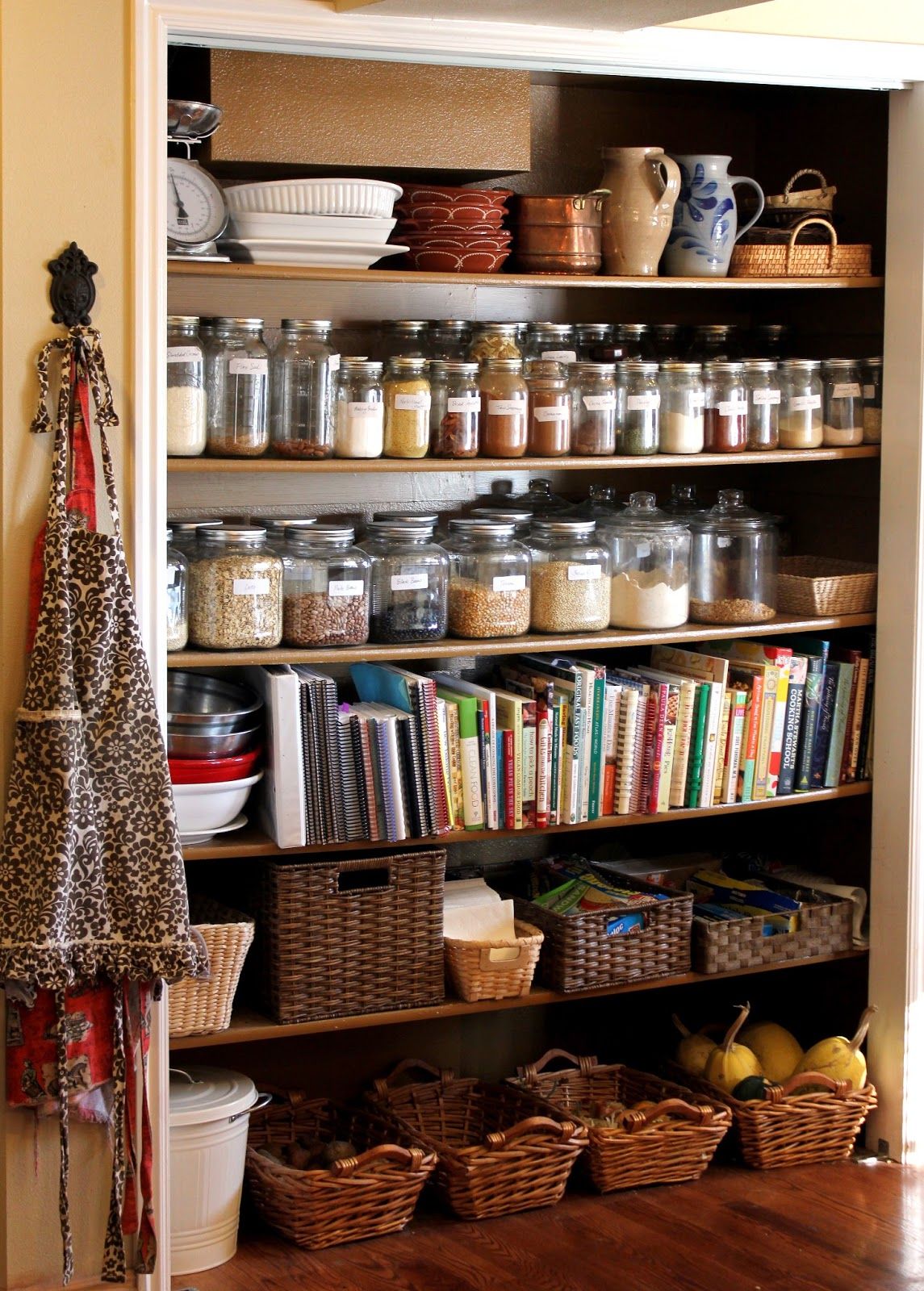 Open-face farmhouse pantry with dark wood shelves lined with labelled mason jars, large glass canisters, a full row of cookbooks, wicker baskets for vegetables, and a printed apron hanging on the side wall