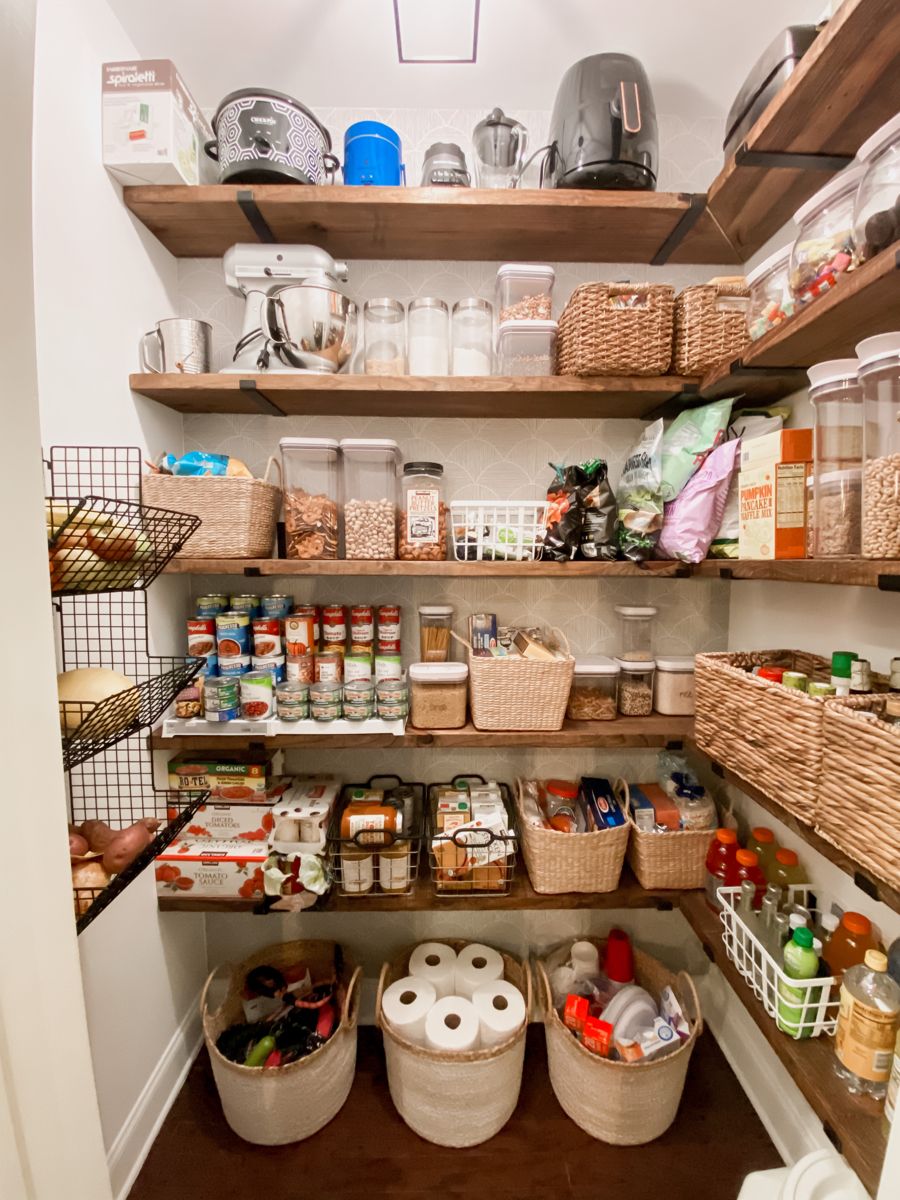 Walk-in farmhouse pantry with dark wood floating shelves and black brackets, organised with wicker baskets, wire bins, OXO containers, a KitchenAid stand mixer, canned goods on a tiered rack, and large floor baskets for bulk storage