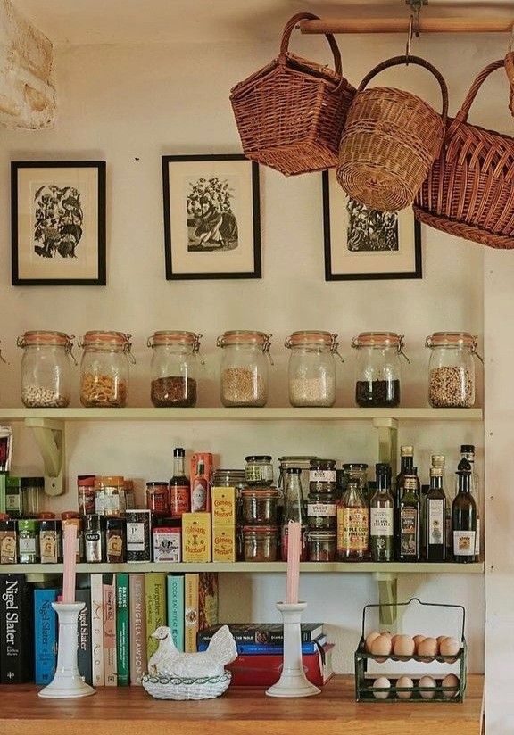 Farmhouse-style open kitchen shelves with rows of swing-top glass jars, bottles of oils and sauces, stacked cookbooks, three framed botanical prints above, and oversized wicker baskets suspended from a ceiling rail