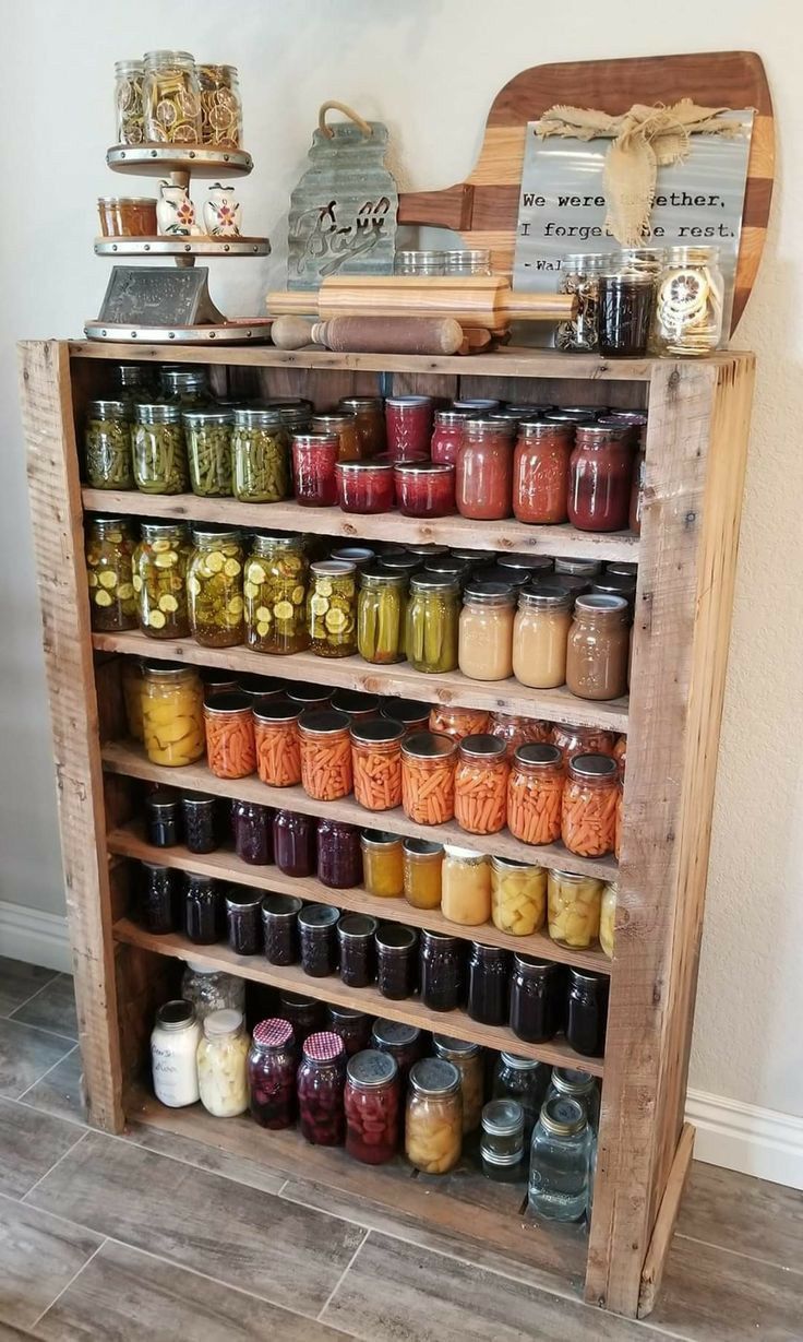Rustic reclaimed pallet wood freestanding shelf unit filled with rows of home-canned mason jars of preserves and pickles in vibrant colours including green, yellow, orange, red, and purple, with a vintage scale and rolling pin on top