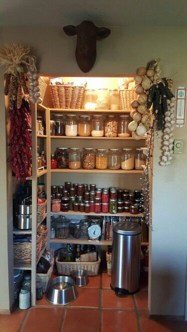 Farmhouse pantry alcove with natural wood shelves holding glass storage jars and home-canned goods, with braids of dried garlic, strings of dried red chillies, and dried herb bundles hanging from the shelf sides and walls