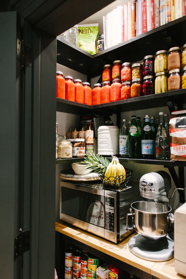 Dark slate-painted farmhouse pantry with open shelving, rows of home-canned tomatoes and colourful preserves, sparkling water bottles, cookbooks on the top shelf, and a built-in butcher block countertop with a KitchenAid mixer and microwave