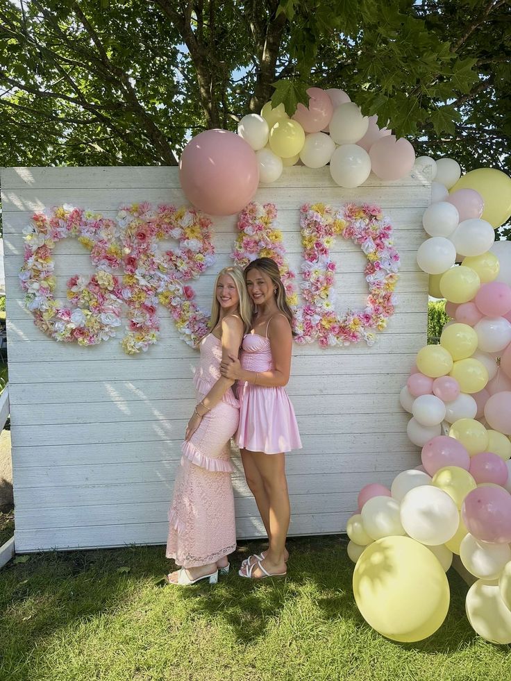 Two girls standing in front of a white wood panel backdrop with large GRAD letters covered in pink yellow and white silk flowers with a pastel balloon garland