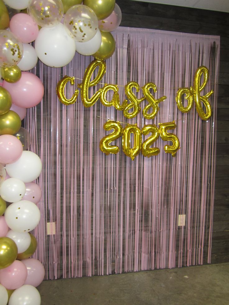 Shimmery pink tinsel fringe curtain graduation backdrop with gold foil script balloons spelling Class of 2025 and a partial balloon garland on the left side