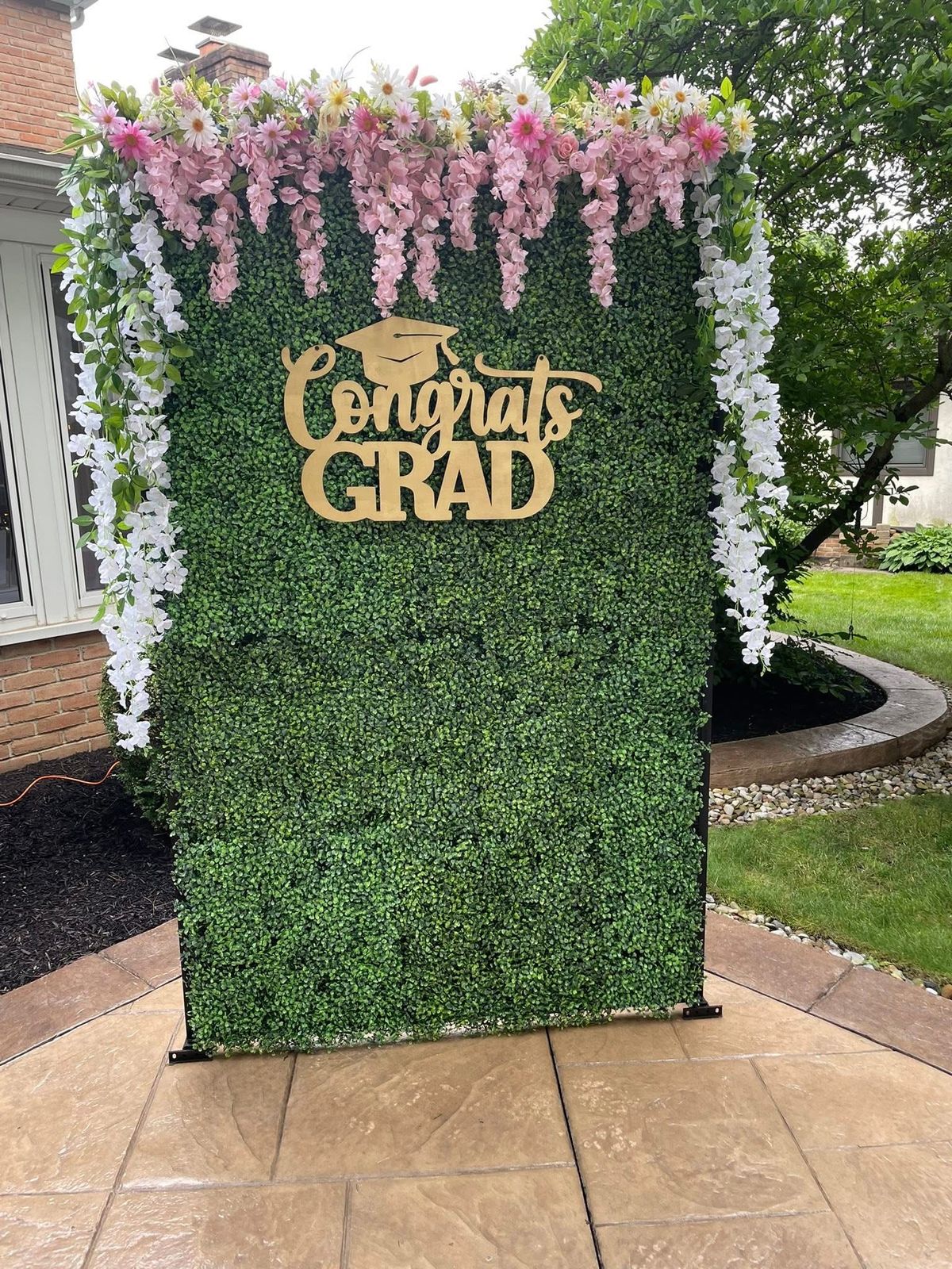 Green boxwood hedge wall backdrop with cascading pink and white faux wisteria on the sides and a gold laser cut Congrats Grad sign mounted in the centre