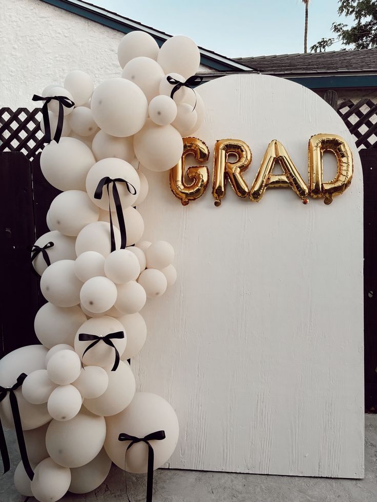 White arched backdrop panel with a cluster of cream balloons decorated with small black ribbon bows on one side and gold foil GRAD letter balloons on the arch face