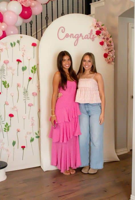 Double white arch panel graduation backdrop with a scattered pink floral pattern on one side and a pink acrylic Congrats sign on the other with a partial balloon and flower arch