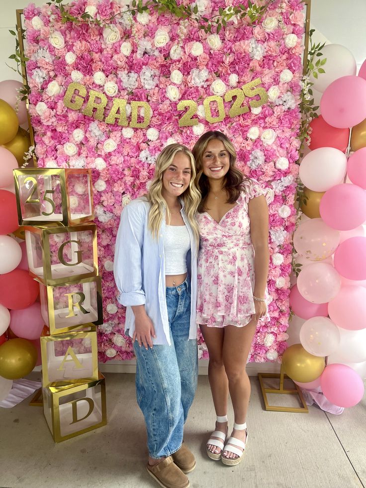 Dense pink and white faux flower wall backdrop with a gold glitter Grad 2025 banner and gold framed acrylic GRAD letter boxes stacked to the side