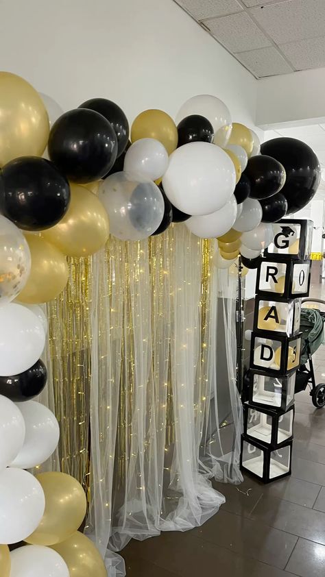 Graduation backdrop with a black white and gold balloon arch over a layered sheer white curtain and gold tinsel backdrop with acrylic GRAD letter boxes to the side