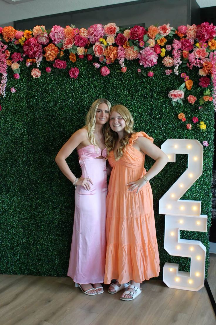 Two girls posing in front of a green boxwood hedge wall topped with vibrant pink orange and coral flowers beside an illuminated marquee number