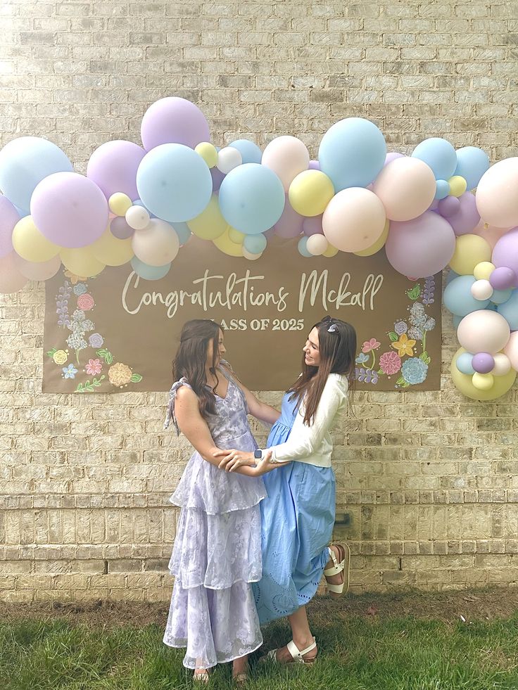 Personalised kraft paper banner reading Congratulations and the grad's name class of 2025 with hand-drawn florals leaned against an exposed brick wall with a pastel balloon garland in lilac blue lemon and blush arching above