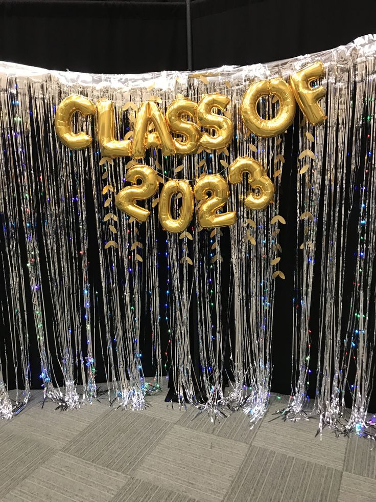 Silver tinsel fringe curtain graduation backdrop with fairy lights woven through the strands and gold foil balloons spelling Class of 2023 positioned in front