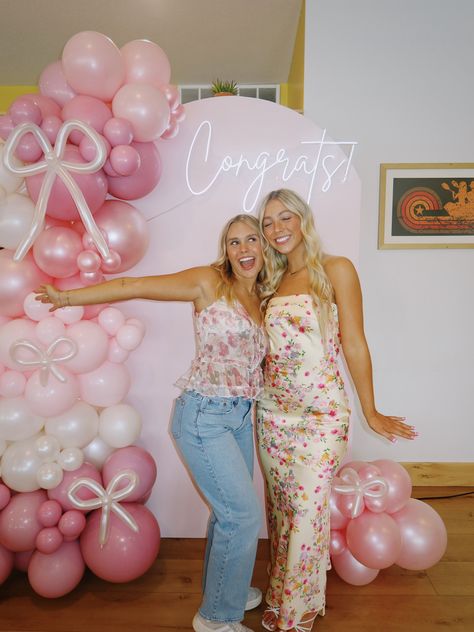 Pale pink arch panel graduation backdrop with a glowing neon Congrats sign at the top and a pink balloon garland featuring oversized bow-shaped balloons in blush pink and mauve