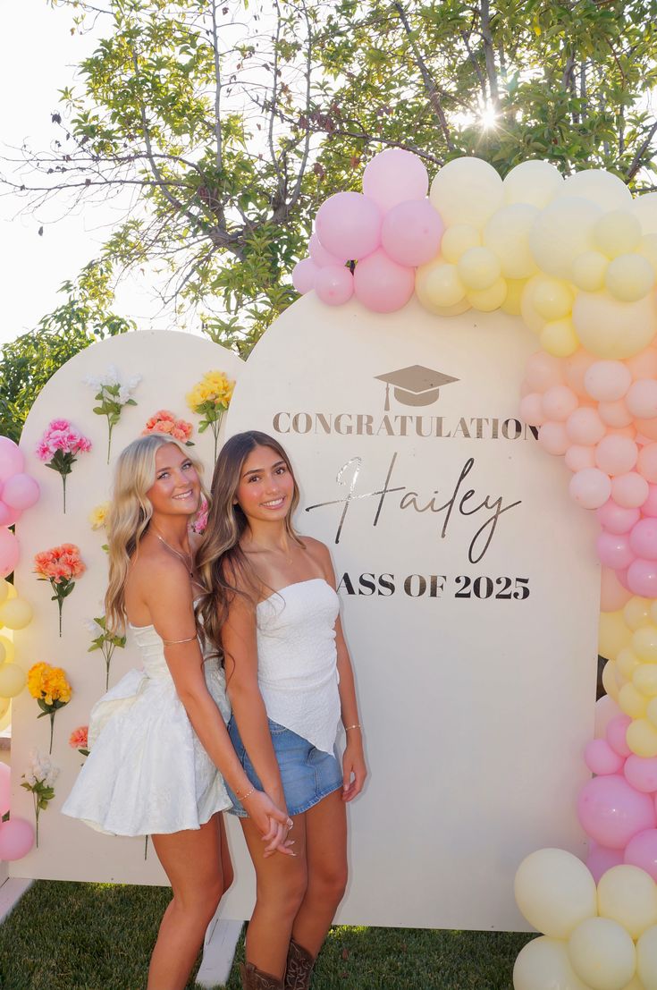 Personalised cream arch graduation sign reading Congratulations and the grad's name class of 2025 surrounded by scattered flowers and a pastel pink and yellow balloon garland
