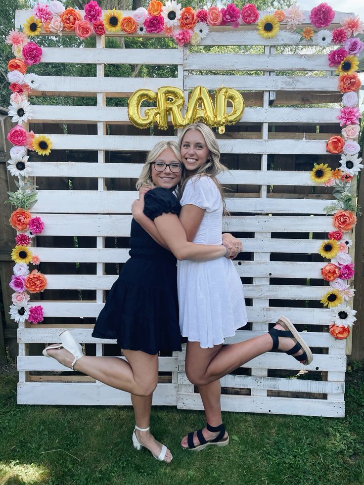 White painted wooden pallet wall decorated with a colourful flower border of sunflowers peonies and roses with gold foil GRAD letter balloons hanging in the centre