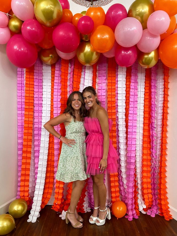 Vibrant graduation party backdrop with layered pink, orange, and white crepe paper fringe curtain topped with a bold balloon arch in matching colours and gold chrome balloons