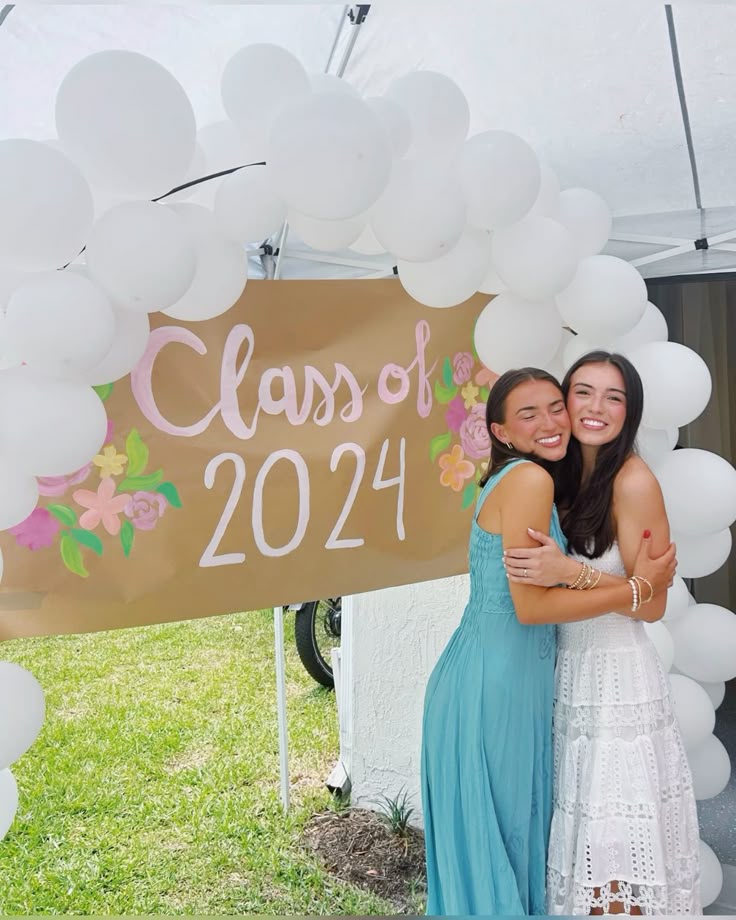 Hand-painted Class of 2024 sign on gold poster board with soft floral details surrounded by a large white balloon arch outdoors