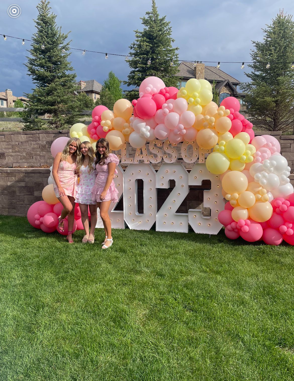 Three girls posing in front of large illuminated marquee numbers reading Class of 2023 surrounded by a massive organic balloon cloud in pink peach yellow and white