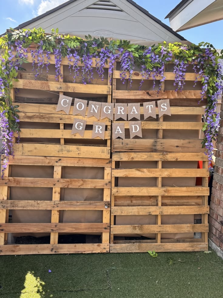 Natural timber pallet wall backdrop with cascading purple faux wisteria garlands draped over the top and a Congrats Grad burlap banner hung across the middle